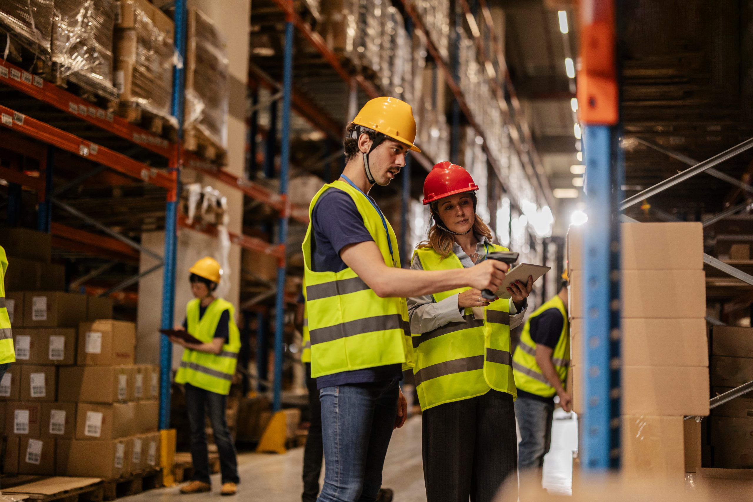 two workers in hard hats and yellow vests scanning boxes in a warehouse