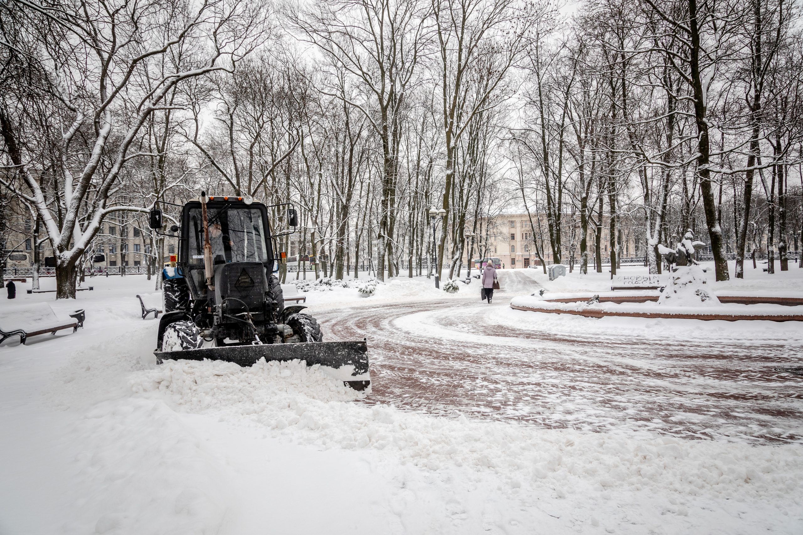 snow plow on roundabout surrounded by snow-covered trees with buildings in the background
