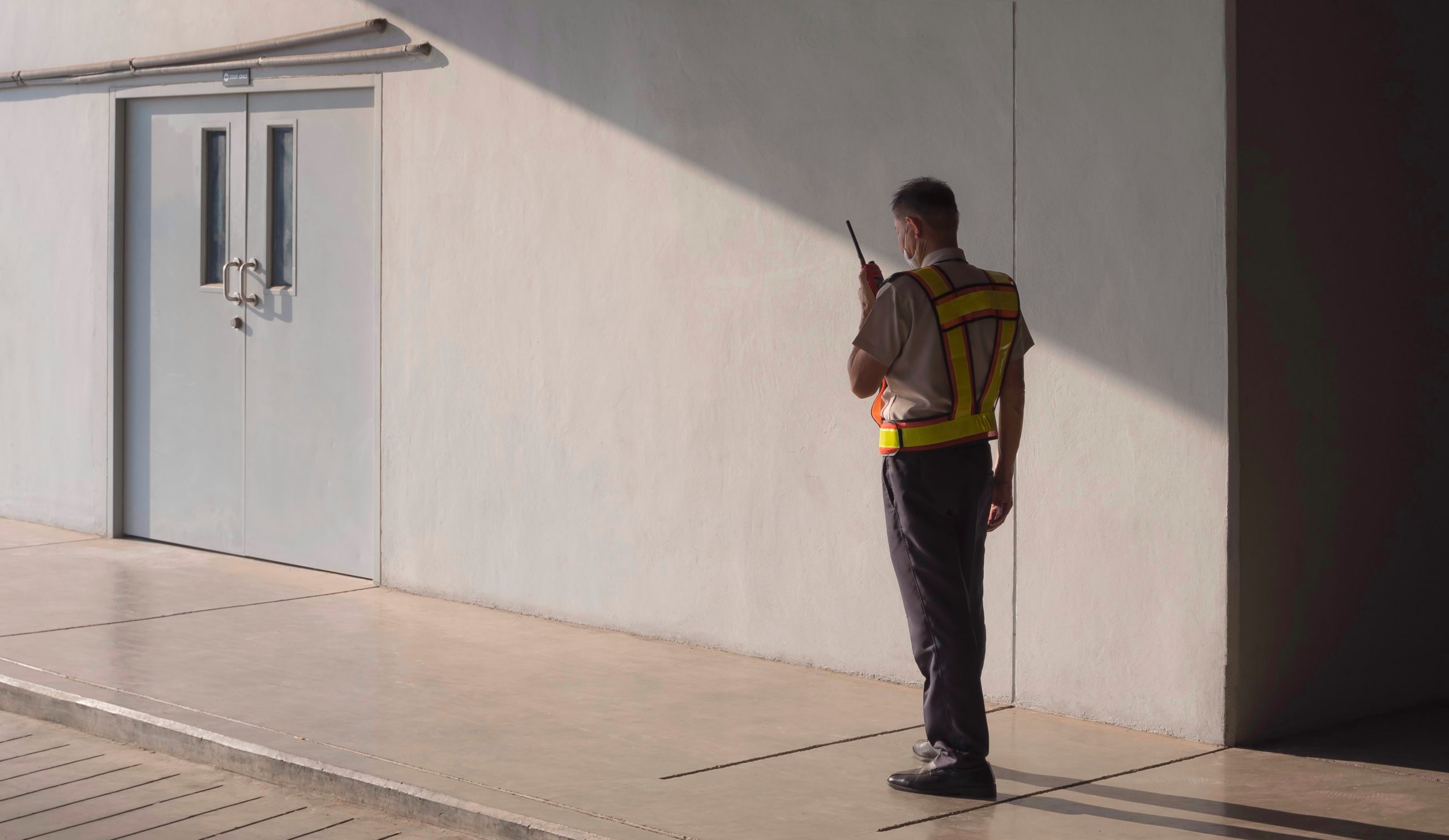 Security guard wearing yellow and orange vest standing outside of a double door and talking on a walkie-talkie
